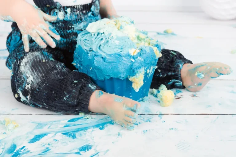 Kindje in donkerblauwe broek tussen de blauwe taartresten op de witte houten vloer, cake smash Zoetermeer
