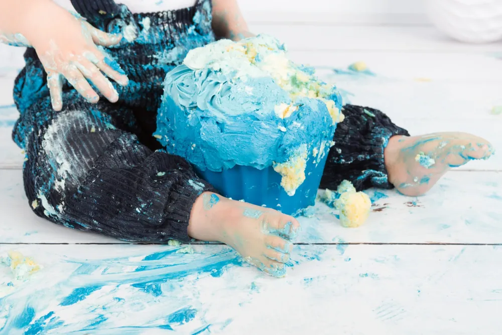 Kindje in donkerblauwe broek tussen de blauwe taartresten op de witte houten vloer, cake smash Zoetermeer